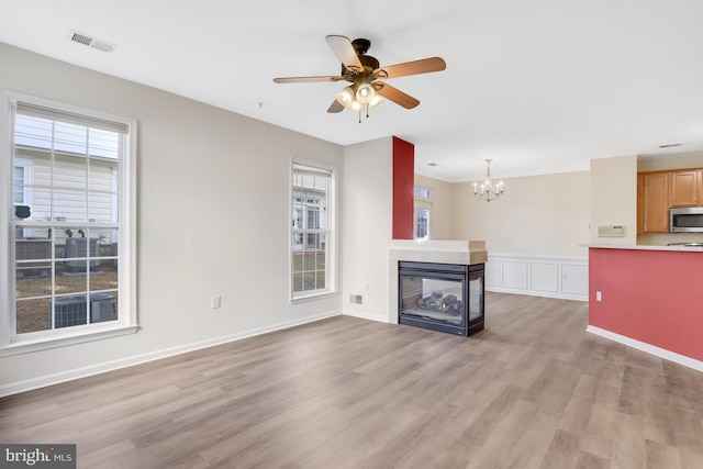 unfurnished living room with ceiling fan with notable chandelier, light hardwood / wood-style flooring, a healthy amount of sunlight, and a multi sided fireplace