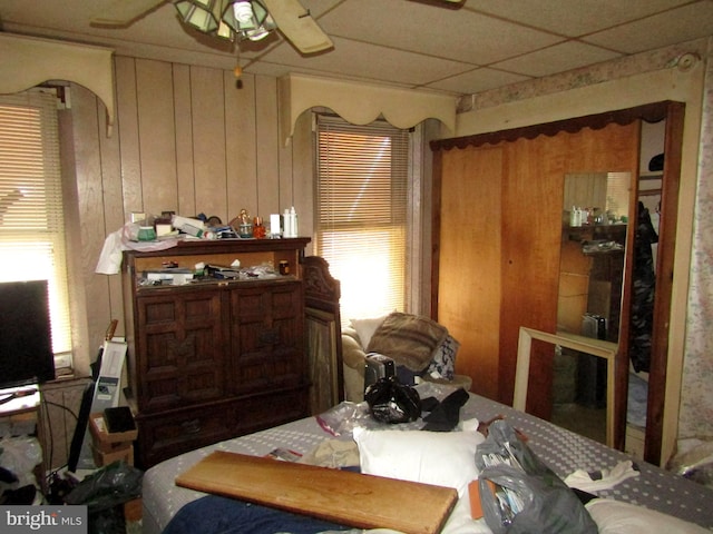 bedroom featuring ceiling fan, wood walls, a drop ceiling, and multiple windows