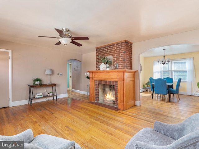 living room with a brick fireplace, ceiling fan with notable chandelier, a baseboard radiator, and light hardwood / wood-style flooring