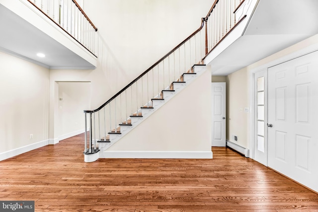 foyer entrance with wood-type flooring, baseboard heating, a towering ceiling, and ornamental molding