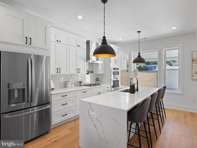 kitchen featuring wall chimney exhaust hood, appliances with stainless steel finishes, a center island with sink, white cabinets, and light wood-type flooring