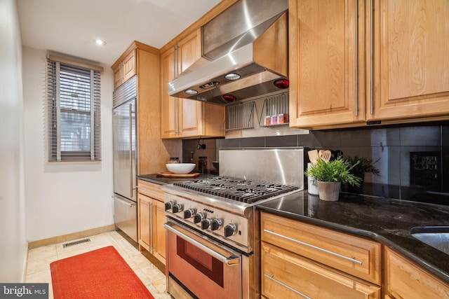 kitchen with backsplash, dark stone counters, wall chimney exhaust hood, high end appliances, and light tile patterned flooring