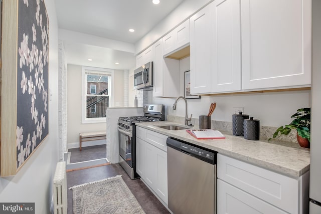 kitchen with sink, radiator heating unit, stainless steel appliances, light stone counters, and white cabinets