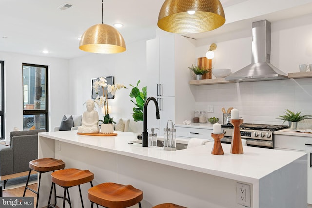 kitchen with a center island with sink, wall chimney range hood, hanging light fixtures, stainless steel gas stove, and tasteful backsplash