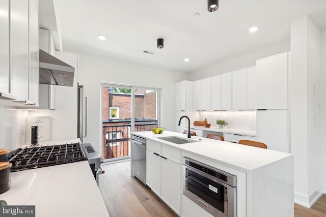 kitchen featuring white cabinetry, sink, stainless steel appliances, a center island with sink, and light wood-type flooring