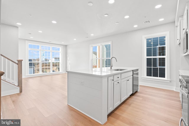 kitchen with light wood-type flooring, a wealth of natural light, sink, white cabinetry, and an island with sink