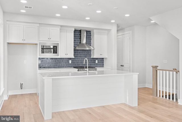 kitchen featuring light wood-type flooring, black microwave, wall chimney range hood, white cabinets, and an island with sink