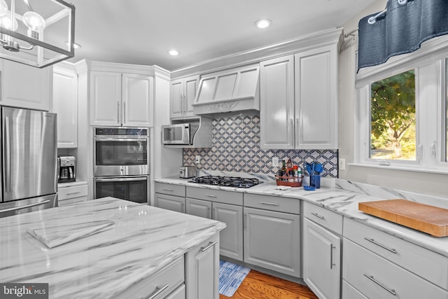 kitchen featuring light stone countertops, light wood-type flooring, custom range hood, stainless steel appliances, and decorative light fixtures