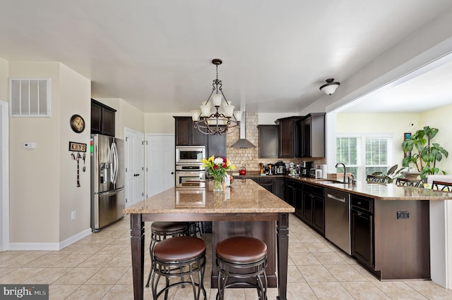 kitchen with hanging light fixtures, stainless steel appliances, wall chimney range hood, an inviting chandelier, and a kitchen island with sink