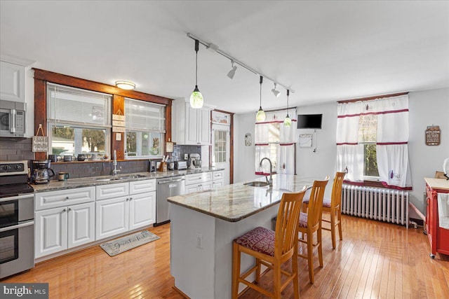 kitchen featuring radiator, white cabinetry, stainless steel appliances, an island with sink, and decorative backsplash