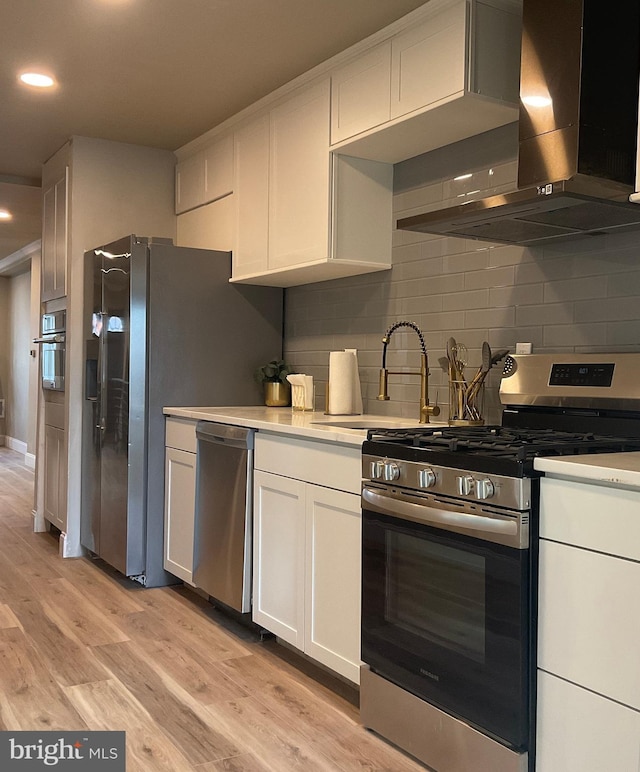 kitchen with appliances with stainless steel finishes, backsplash, wall chimney range hood, light hardwood / wood-style floors, and white cabinetry