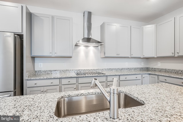 kitchen with sink, wall chimney exhaust hood, light stone counters, stainless steel fridge, and black electric cooktop