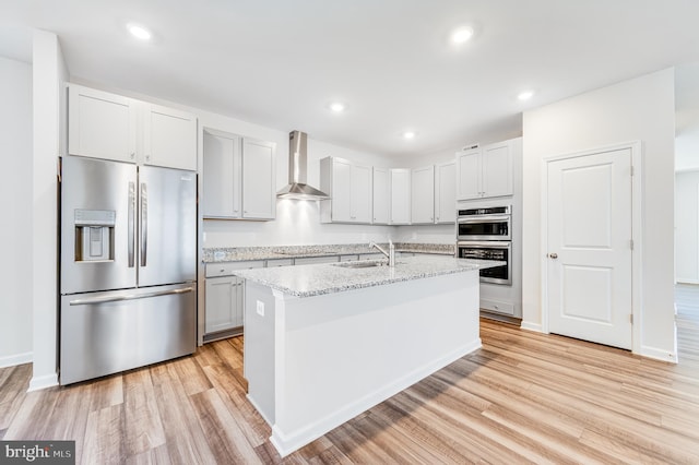 kitchen featuring a center island with sink, light wood-type flooring, wall chimney range hood, and appliances with stainless steel finishes