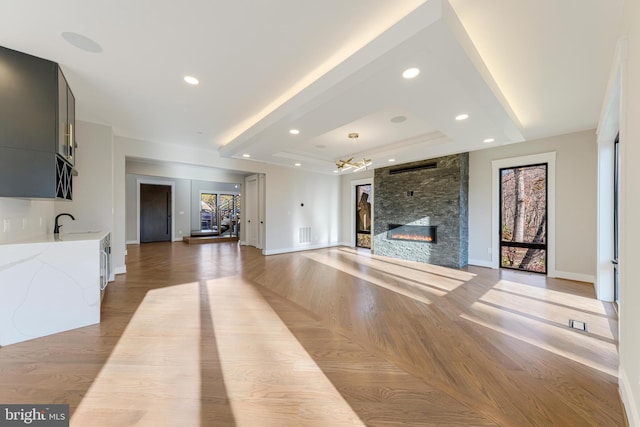 unfurnished living room featuring sink, a raised ceiling, a stone fireplace, a chandelier, and light wood-type flooring