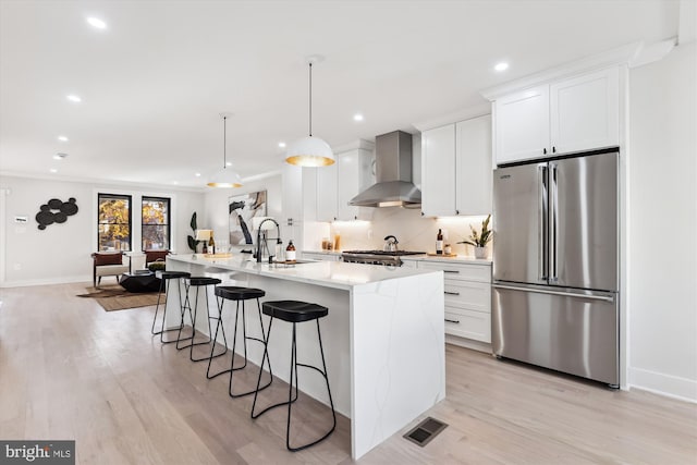 kitchen featuring a kitchen island with sink, hanging light fixtures, wall chimney exhaust hood, appliances with stainless steel finishes, and white cabinetry