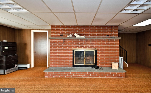 unfurnished living room featuring carpet, a drop ceiling, wooden walls, and a brick fireplace