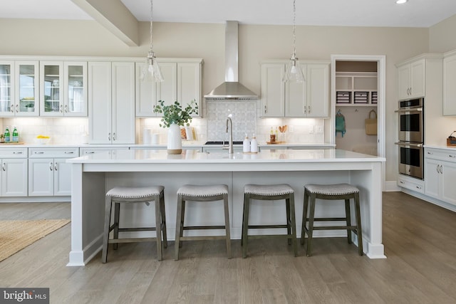 kitchen featuring white cabinetry, wall chimney range hood, a kitchen island with sink, and double oven