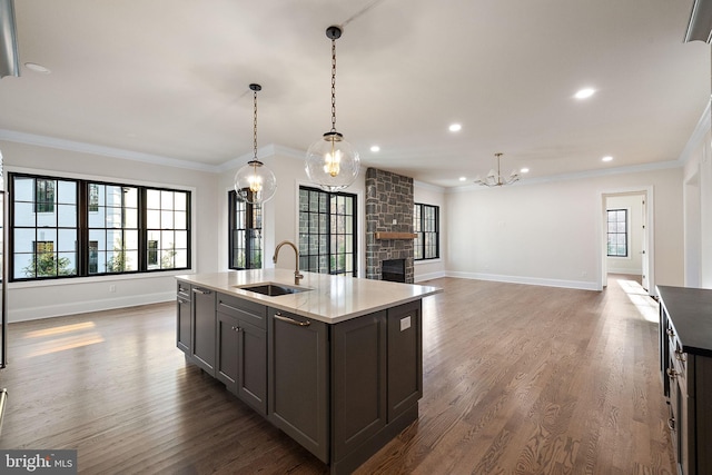 kitchen with dark wood-type flooring, a stone fireplace, sink, an island with sink, and decorative light fixtures