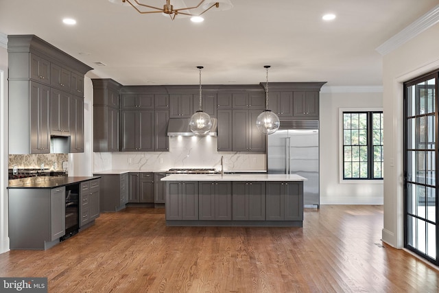 kitchen with hanging light fixtures, gray cabinets, ornamental molding, an island with sink, and stainless steel built in refrigerator