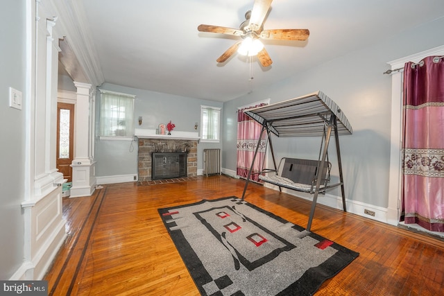 exercise room with wood-type flooring, a stone fireplace, ceiling fan, and radiator