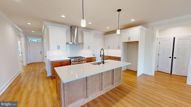 kitchen with sink, wall chimney exhaust hood, stainless steel stove, a center island with sink, and white cabinets