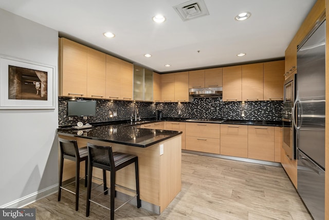 kitchen with stainless steel appliances, a kitchen breakfast bar, dark stone countertops, light hardwood / wood-style floors, and light brown cabinetry