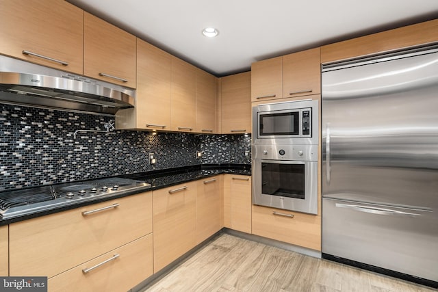 kitchen featuring built in appliances, light brown cabinets, dark stone countertops, and backsplash