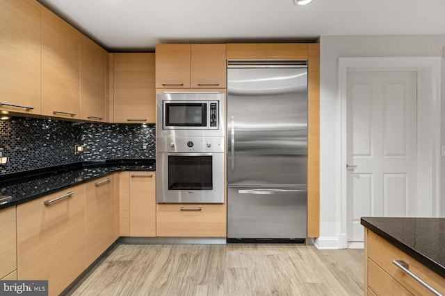 kitchen featuring light brown cabinets, tasteful backsplash, built in appliances, dark stone counters, and light hardwood / wood-style floors