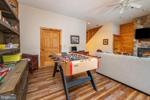 recreation room featuring ceiling fan, a stone fireplace, wood walls, wood-type flooring, and a textured ceiling