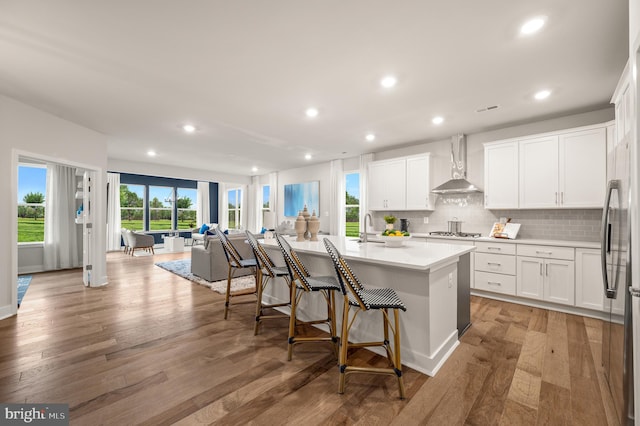 kitchen with white cabinets, light wood-type flooring, an island with sink, and wall chimney range hood