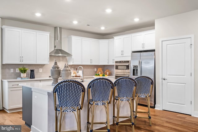 kitchen with white cabinets, a center island with sink, wall chimney exhaust hood, light wood-type flooring, and appliances with stainless steel finishes