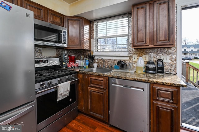 kitchen with appliances with stainless steel finishes, dark hardwood / wood-style flooring, tasteful backsplash, dark stone counters, and sink