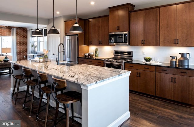 kitchen with decorative light fixtures, stainless steel appliances, dark wood-type flooring, and an island with sink