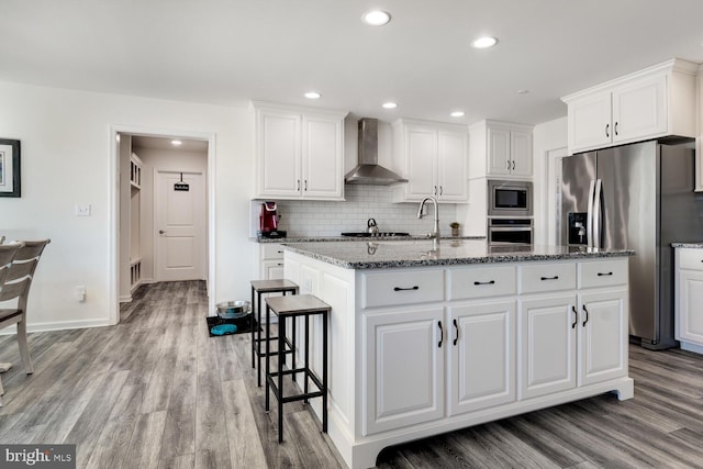 kitchen with wall chimney exhaust hood, light hardwood / wood-style flooring, a center island with sink, white cabinets, and appliances with stainless steel finishes