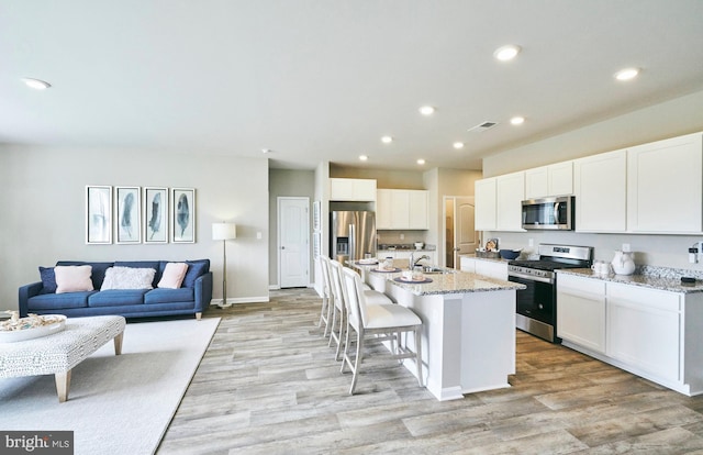 kitchen with appliances with stainless steel finishes, light wood-type flooring, light stone counters, a kitchen island with sink, and white cabinetry