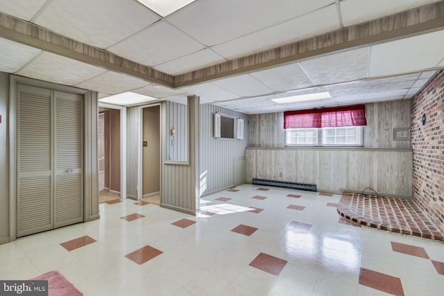 basement featuring a paneled ceiling, wood walls, a wall mounted air conditioner, and a baseboard radiator