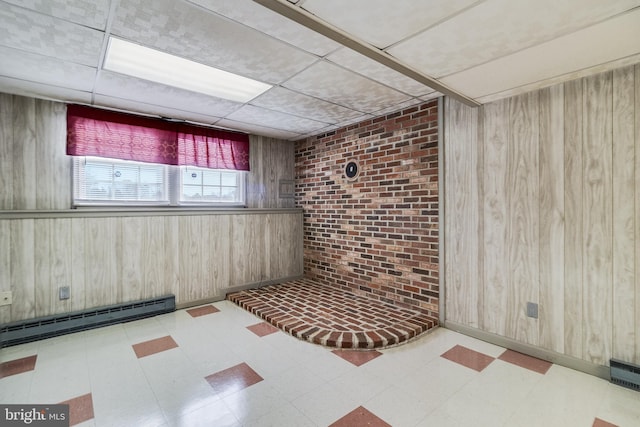 basement featuring brick wall, a paneled ceiling, a baseboard heating unit, and wood walls