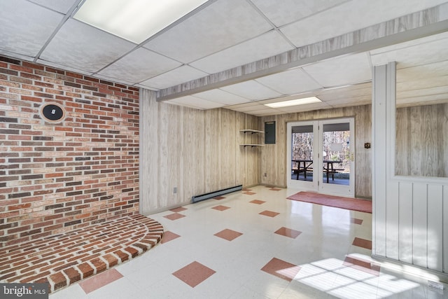 basement featuring a paneled ceiling, baseboard heating, wooden walls, and french doors