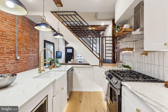 kitchen featuring wall chimney range hood, light wood-type flooring, appliances with stainless steel finishes, white cabinetry, and brick wall