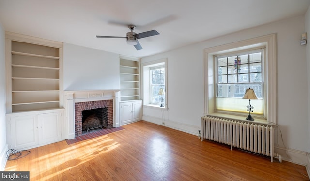 unfurnished living room with a brick fireplace, radiator, built in shelves, ceiling fan, and hardwood / wood-style floors