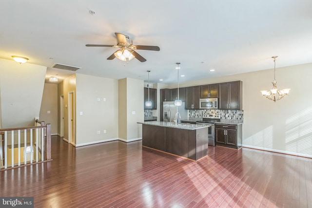 kitchen with dark wood-type flooring, a center island with sink, hanging light fixtures, appliances with stainless steel finishes, and tasteful backsplash