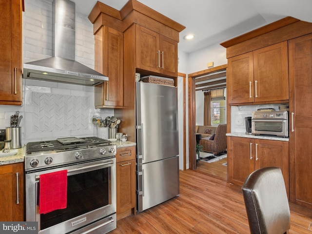 kitchen featuring light stone countertops, appliances with stainless steel finishes, light wood-type flooring, decorative backsplash, and wall chimney range hood