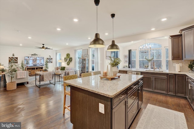 kitchen with dark hardwood / wood-style flooring, light stone counters, ceiling fan with notable chandelier, a kitchen island, and hanging light fixtures