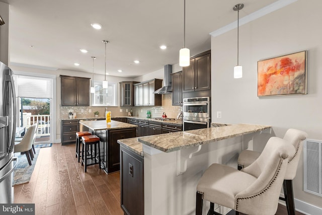 kitchen featuring wall chimney exhaust hood, dark hardwood / wood-style floors, decorative light fixtures, a kitchen bar, and dark brown cabinetry