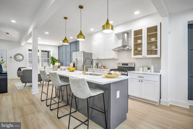 kitchen featuring a center island with sink, white cabinets, wall chimney range hood, hanging light fixtures, and appliances with stainless steel finishes