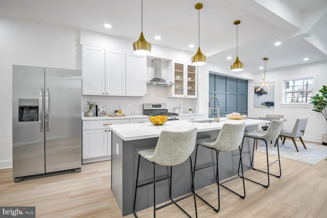 kitchen with wall chimney exhaust hood, stainless steel appliances, decorative light fixtures, a kitchen island with sink, and white cabinets