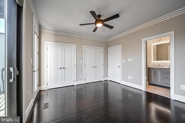 unfurnished bedroom with multiple closets, ceiling fan, ensuite bath, dark wood-type flooring, and crown molding