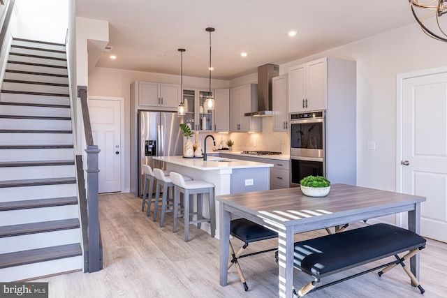 kitchen featuring hanging light fixtures, wall chimney range hood, an island with sink, gray cabinets, and appliances with stainless steel finishes