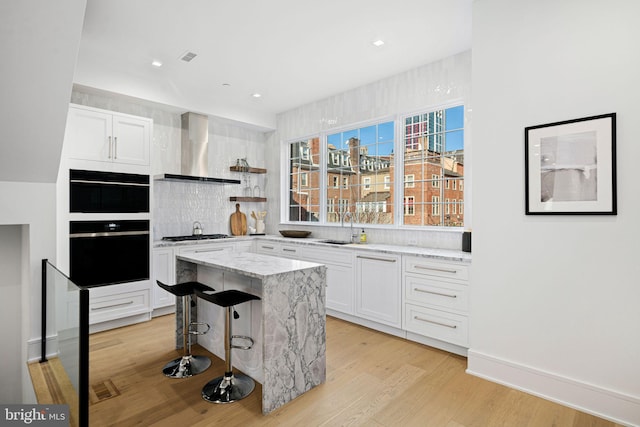 kitchen featuring wall chimney range hood, a kitchen island, light hardwood / wood-style floors, a kitchen bar, and white cabinetry