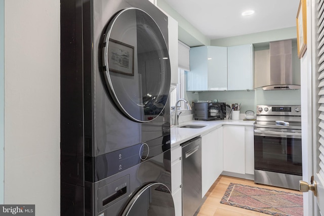 kitchen with wall chimney exhaust hood, stainless steel appliances, light hardwood / wood-style flooring, stacked washer / dryer, and white cabinetry